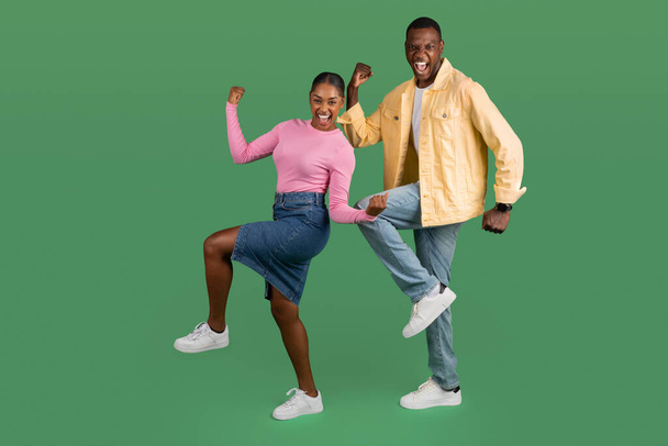 Emotional happy millennial black man and woman wearing casual outfits clenching fists, exclaiming and dancing, couple celebrating success isolated on green studio background, copy space - Photo, Image