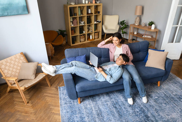 Японские супруги наслаждаются просмотром видео онлайн на ноутбуке, расслабляясь на диване в комфортной гостиной дома. Интернет-технологии и семейный компьютерный досуг - Фото, изображение
