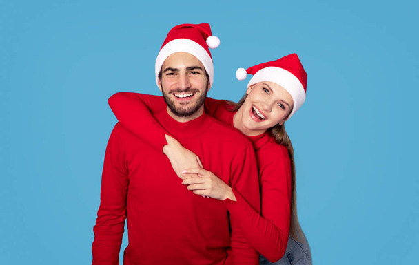 Buone feste. Allegro sorridente giovane coppia in cappelli di Babbo Natale in posa su sfondo blu, gioioso amorevole uomo e donna che indossa maglioni rossi in posa presso la fotocamera insieme, godendo di umore di Natale - Foto, immagini
