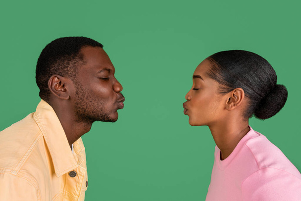 Boční pohled na milujícího mladého černocha a ženu líbající se zavřenýma očima, kopírující prostor mezi nimi, izolovaný na zeleném pozadí studia. Láska, náklonnost, sblížení, vztahy - Fotografie, Obrázek