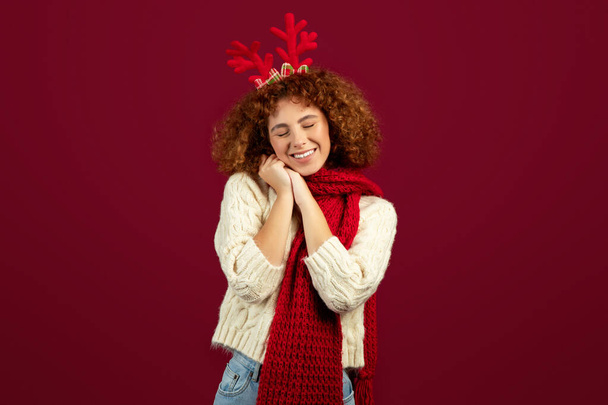 Счастливая женщина-подросток с оленьими рогами в праздничном настроении Рождества и Нового года, наслаждаясь зимними праздниками, изолированная на красном студийном фоне, студия. Сезонная радость, образ жизни и мечта - Фото, изображение
