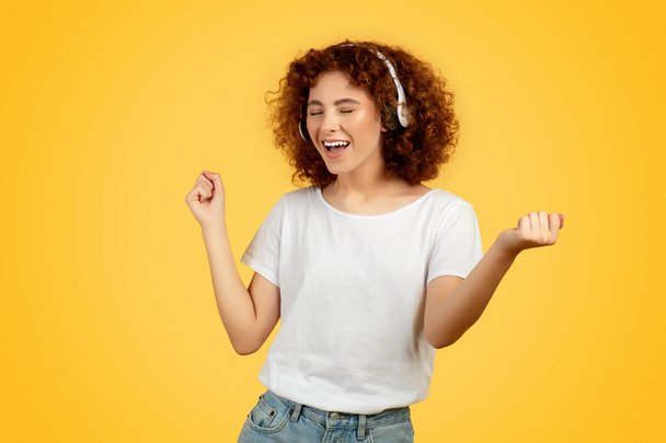 Frohe europäische Teenagerin, auf gelbem Hintergrund, hört Musik über drahtlose Kopfhörer, die Freude und Freiheit ausdrückt. Jugendlicher Überschwang und leidenschaftliche Energie, Tanz und Freizeit - Foto, Bild