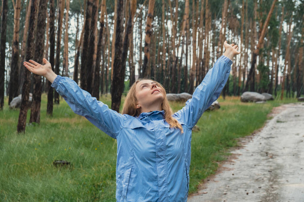 Femme blonde ravie en imperméable bleu profitant du silence vert naturel bois de l'environnement dans la forêt. Joyeux émotion bras ouverts à l'extérieur par temps pluvieux. Unité avec la nature bien-être mental physique  - Photo, image