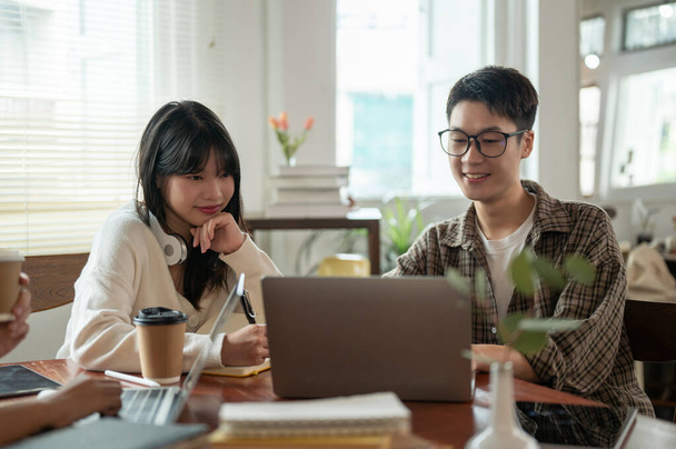 Un joven estudiante asiático inteligente en gafas está trabajando con una amiga, haciendo un co-proyecto en una computadora portátil en una cafetería juntos. Conceptos de amistad y educación - Foto, Imagen