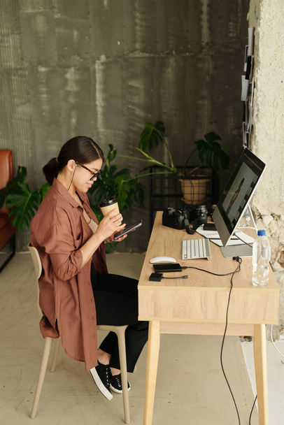 Joven diseñador o fotógrafo mirando a través de nuevas tomas de modelo de moda en el teléfono inteligente mientras está sentado en el lugar de trabajo y tomando café - Foto, imagen