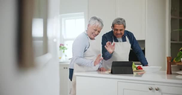 Κουζίνα, χαιρετισμός και ηλικιωμένο ζευγάρι με tablet, βιντεοκλήση και επικοινωνία με σύνδεση, ποδιά και μαγείρεμα. Αρχική σελίδα, ευτυχισμένος ηλικιωμένος άνδρας και ηλικιωμένη γυναίκα με την τεχνολογία, online οδηγίες και κύμα. - Πλάνα, βίντεο