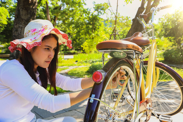 Азиатская женщина в шапке и ботаническом саду на велосипеде с пышными зелеными деревьями и тенистыми деревьями сидит и проверяет давление в шинах, испытывая трудности с ездой, потому что шины слишком слабы. - Фото, изображение