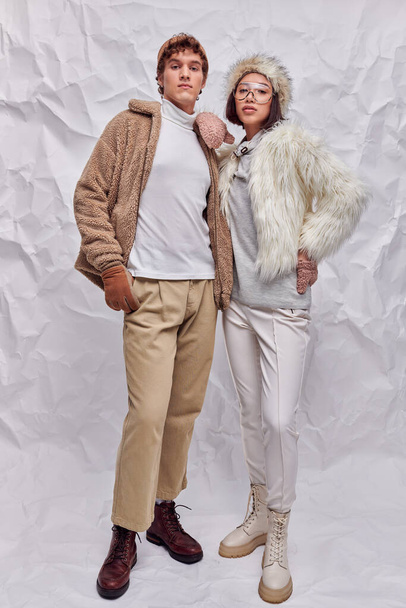 young asian model in warm attire posing with hand on hip near trendy man on white textured backdrop - Photo, Image