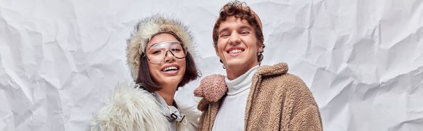 felice coppia interrazziale in elegante abbigliamento invernale guardando la fotocamera su sfondo bianco strutturato, banner - Foto, immagini