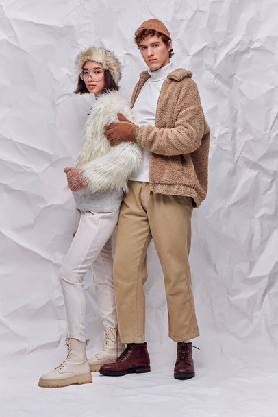 молодий стильний чоловік обіймає азіатську жінку в хутряній куртці та окулярах на збитому паперовому фоні - Фото, зображення