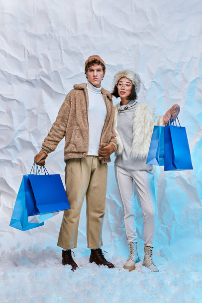 модна міжрасова пара з блакитними сумками на білому снігу в студії, зимові покупки - Фото, зображення