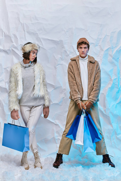 μοντέρνο διαφυλετικό ζευγάρι με μπλε τσάντες ψώνια στέκεται στο χιόνι στο στούντιο, το χειμώνα στυλ - Φωτογραφία, εικόνα