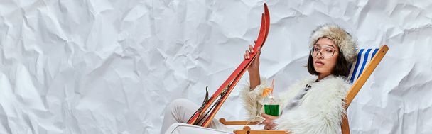 asian woman in winter attire in deck chair with skis and cocktail on white textured backdrop, banner - Photo, Image