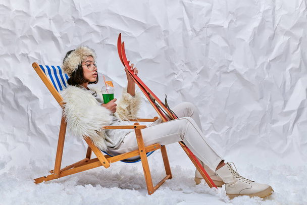 aasialainen nainen lämpimässä trendikäs asu sukset ja cocktail rentouttava kansituoli lumella studiossa - Valokuva, kuva