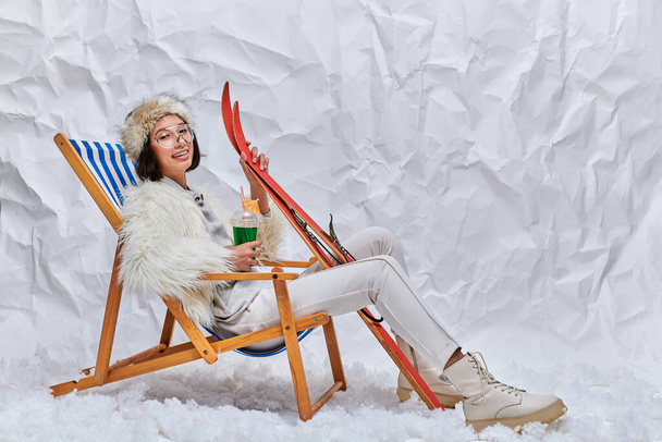 feliz asiático modelo en invierno ropa sentado en cubierta silla con cóctel y esquís en snowy studio - Foto, Imagen