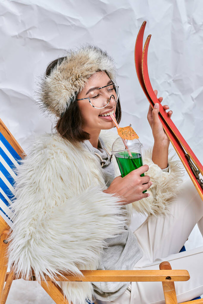 feliz asiático mujer con esquís beber caliente teddy cóctel con cerrado ojos en blanco texturizado telón de fondo - Foto, Imagen