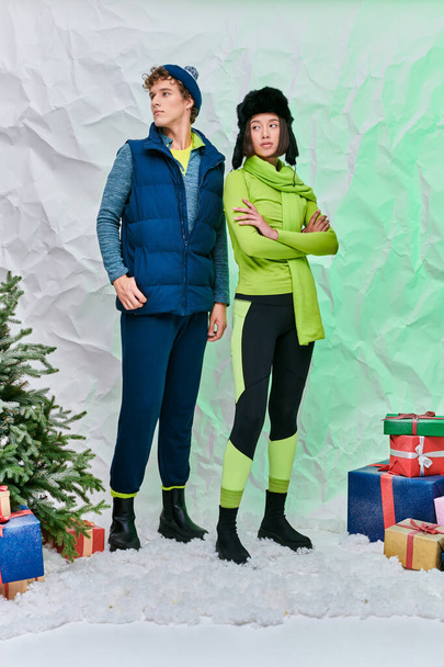 wielonarodowa para w ciepłym ubraniu w pobliżu pudełek prezentów i choinki na śniegu w studio, sezon świąteczny - Zdjęcie, obraz