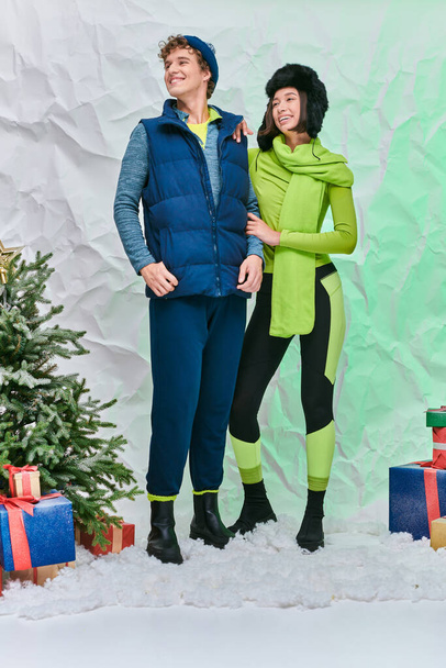 wesołe międzyrasowe modele w ciepłym stroju w pobliżu prezentów i choinki na śniegu w studio - Zdjęcie, obraz