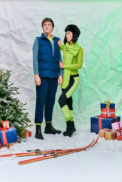 冬のモダンな異人種間のカップルは,スタジオで雪の上のプレゼント,スキー,クリスマスツリーの近くに着用します - 写真・画像