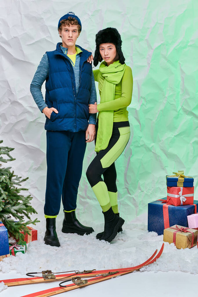 модна міжрасова пара в теплому одязі біля подарунків, лиж і ялинки в засніженій студії - Фото, зображення