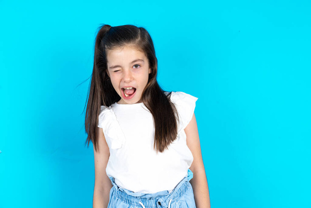 Beautiful caucasian kid girl standing over blue studio background winking looking at the camera with sexy expression, cheerful and happy face. - Photo, Image