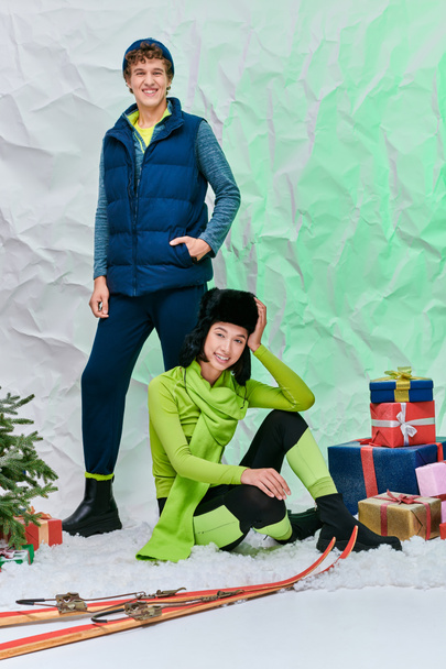 pulzující mezirasový pár pózující na sněhu ve studiu v blízkosti vánoční strom, dárkové krabice a lyže - Fotografie, Obrázek