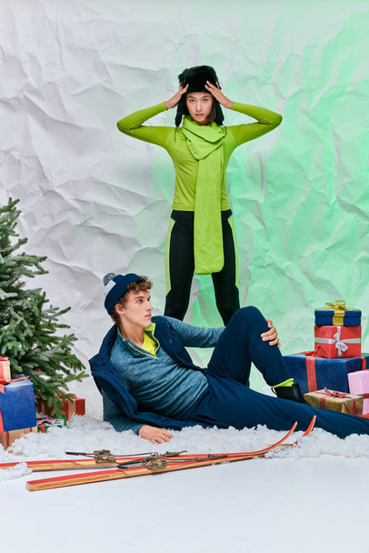 κομψό ασιατικό μοντέλο κοιτάζοντας κάμερα κοντά στον άνθρωπο στο χιόνι κοντά σε κουτιά δώρων και χριστουγεννιάτικο δέντρο στο στούντιο - Φωτογραφία, εικόνα