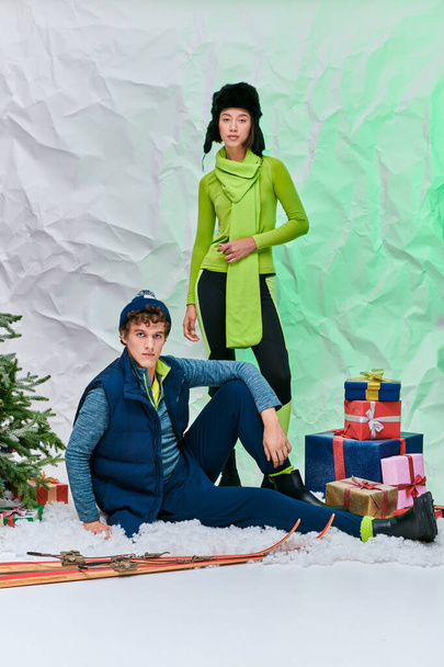 модная многонациональная пара, смотрящая в камеру рядом с подарками и елкой в снежной студии - Фото, изображение