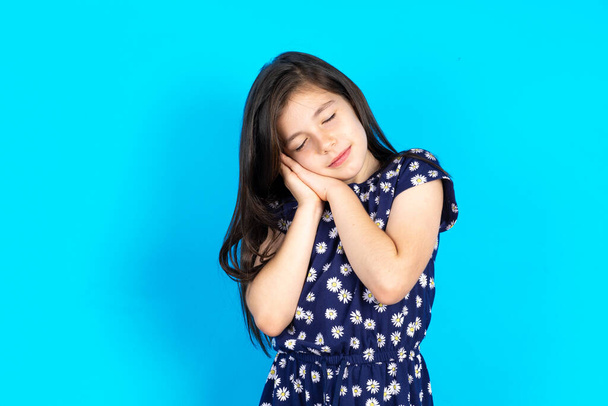 Belle jeune fille brune caucasienne portant une robe sur fond bleu avec les yeux fermés s'appuyant sur les paumes faisant geste de sommeil. - Photo, image