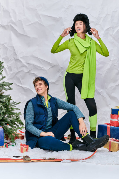 веселая многонациональная пара, смотрящая в заснеженной студии возле рождественской елки и подарочных коробок - Фото, изображение