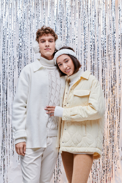 joyful interracial couple in white jackets standing near silver tinsel on backdrop, christmas spirit - Photo, Image