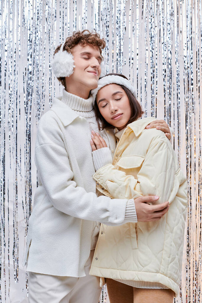 happy interracial couple in cozy winter wear embracing with closed eyes near shiny tinsel in studio - Photo, Image