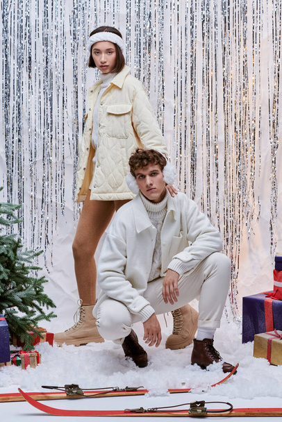modelos interracial en ropa de invierno posando cerca de árbol de Navidad, regalos y decoración brillante en el estudio - Foto, imagen