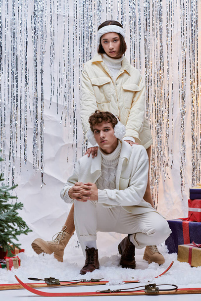 многонациональная пара в уютной одежде, смотрящая в камеру возле рождественской елки, подарков и серебряной мишуры - Фото, изображение