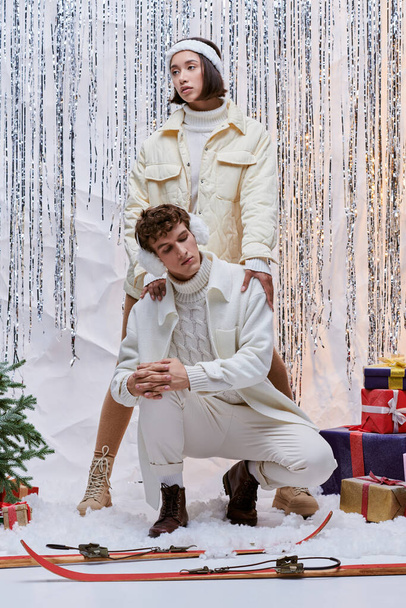 Multiethnische Models in trendiger Winterkleidung posieren in der Nähe von Weihnachtsbaum, Geschenkschachteln und silbernem Lametta - Foto, Bild