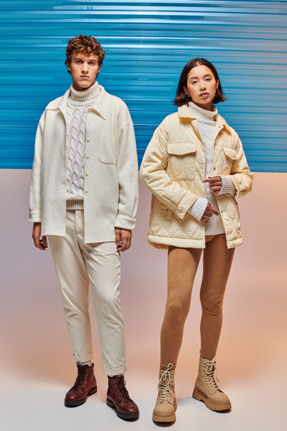 pełna długość międzyrasowej pary w ciepłych kurtkach i skórzanych butach pozujących w pobliżu niebieskiego plastikowego arkusza - Zdjęcie, obraz