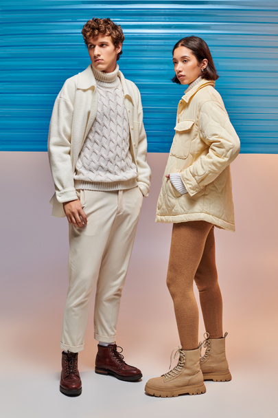 coppia interrazziale in abbigliamento invernale alla moda posa vicino foglio di plastica blu, moda stagionale - Foto, immagini