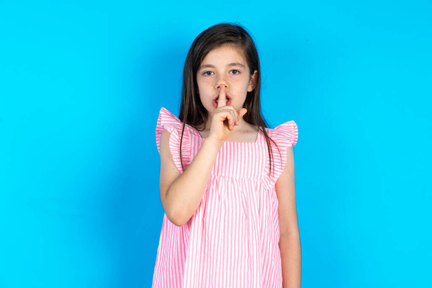 Mooi meisje draagt roze jurk over blauwe achtergrond maakt stilte gebaar, houdt de vinger over de lippen. Stilte en geheim concept. - Foto, afbeelding
