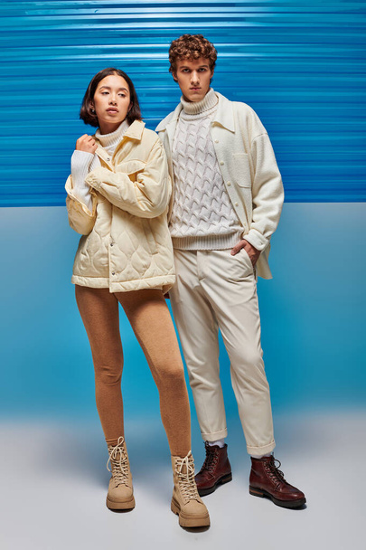 trendy man with hand in pocket near asian woman in warm clothes on blue backdrop with plastic panel - Photo, Image