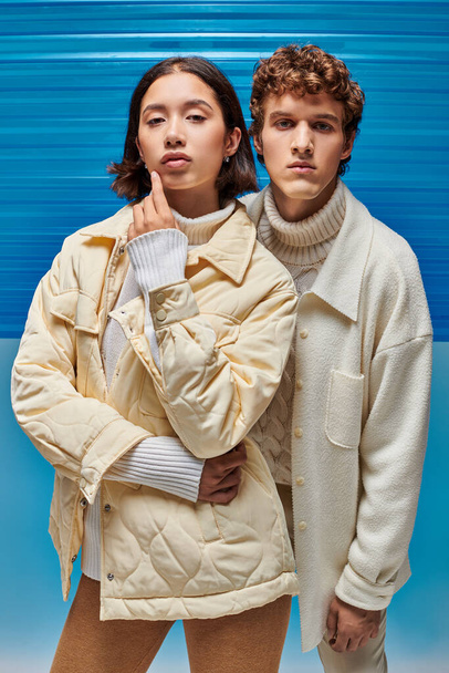 winter mode campagne, interraciale paar jassen op zoek naar camera op blauwe plastic achtergrond - Foto, afbeelding
