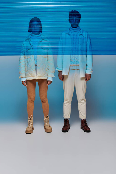 coppia interrazziale in giacche e stivali di pelle in piedi dietro pannello di plastica blu, stile invernale - Foto, immagini