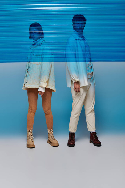 pareja multiétnica en chaquetas y botas de cuero posando detrás de lámina de plástico azul, moda de invierno - Foto, Imagen