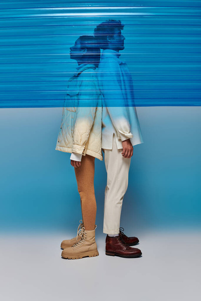 vista lateral do casal interracial em roupas quentes de pé de volta para trás atrás de plástico azul em estúdio - Foto, Imagem