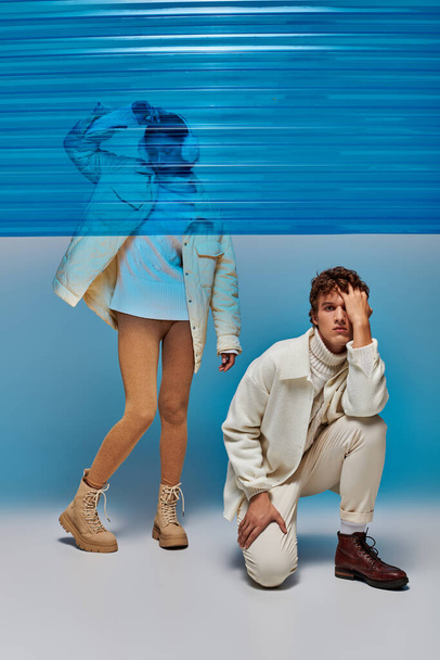 man in trendy kleding op zoek naar camera in de buurt van aziatische model achter blauw plastic blad, winter trend - Foto, afbeelding