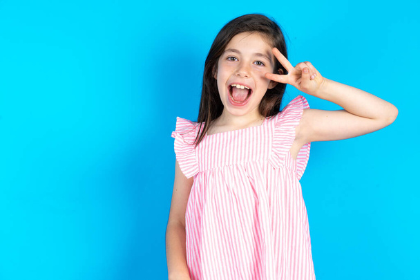 mooi kind meisje dragen roze jurk over blauwe achtergrond doen vrede symbool met vingers over gezicht glimlachend vrolijk tonen overwinning - Foto, afbeelding