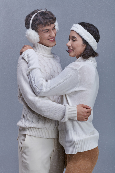 happy multiethnic couple in warm sweaters embracing under falling snowflakes on grey background - Photo, Image