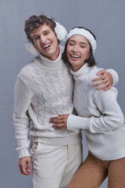 podekscytowana para międzyrasowa w ciepłych swetrach obejmująca i śmiejąca się pod spadającym śniegiem, szczęśliwa zima - Zdjęcie, obraz