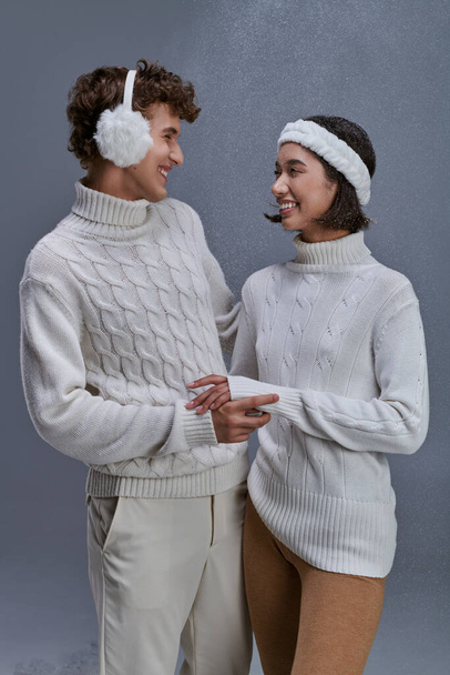 ευτυχισμένο διαφυλετικό ζευγάρι σε πουλόβερ κοιτάζοντας ο ένας τον άλλον σε γκρι χιονισμένο φόντο, ρομαντικό χειμώνα - Φωτογραφία, εικόνα