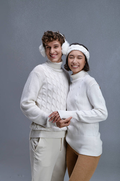 cheerful multiethnic couple in sweaters smiling under falling snow on grey backdrop, trendy winter - Photo, Image