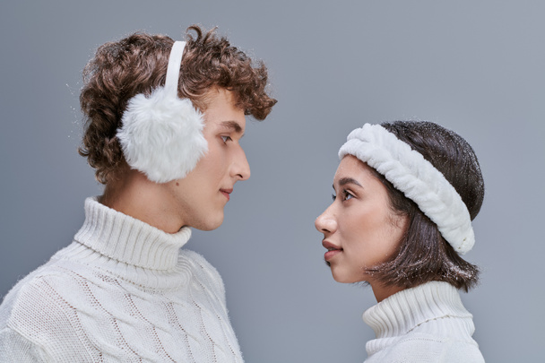 side view of interracial couple in white sweaters and with snowy hair looking at each other on grey - Photo, Image